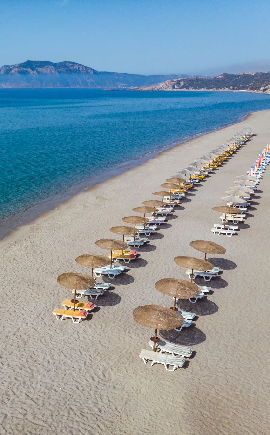 Kardamena beach in Kos, Greece, with rows of empty sun loungers and straw umbrellas on sandy shore.