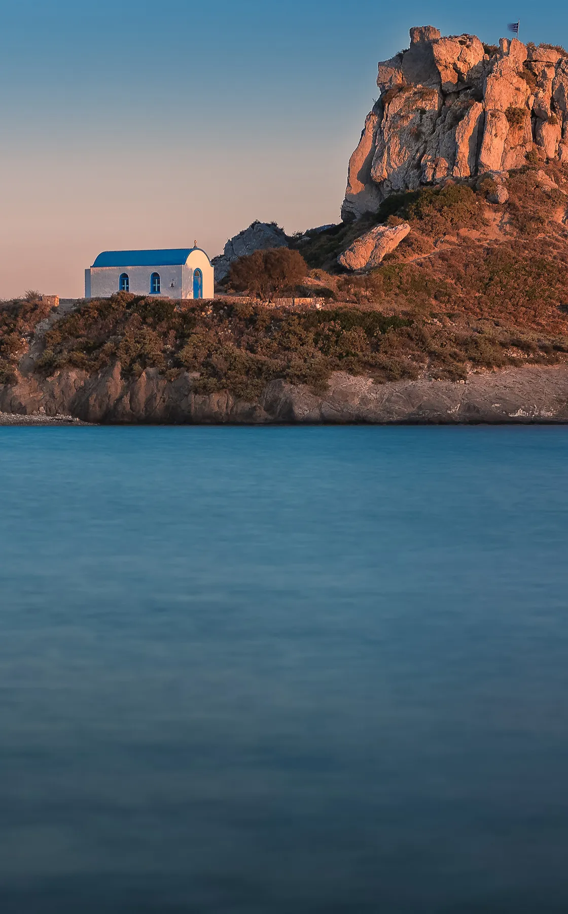 Small chapel with a blue roof near rocky hill and calm sea in Kefalos, Kos, Greece at sunset.