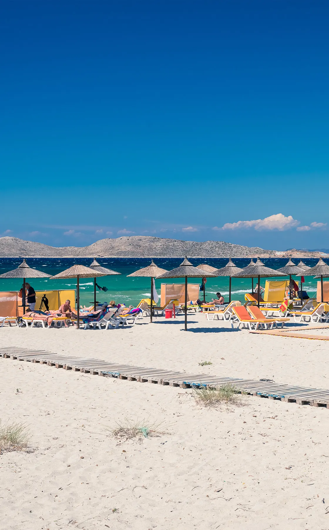 Sandy beach with sun loungers and straw umbrellas near a turquoise sea under a clear blue sky.