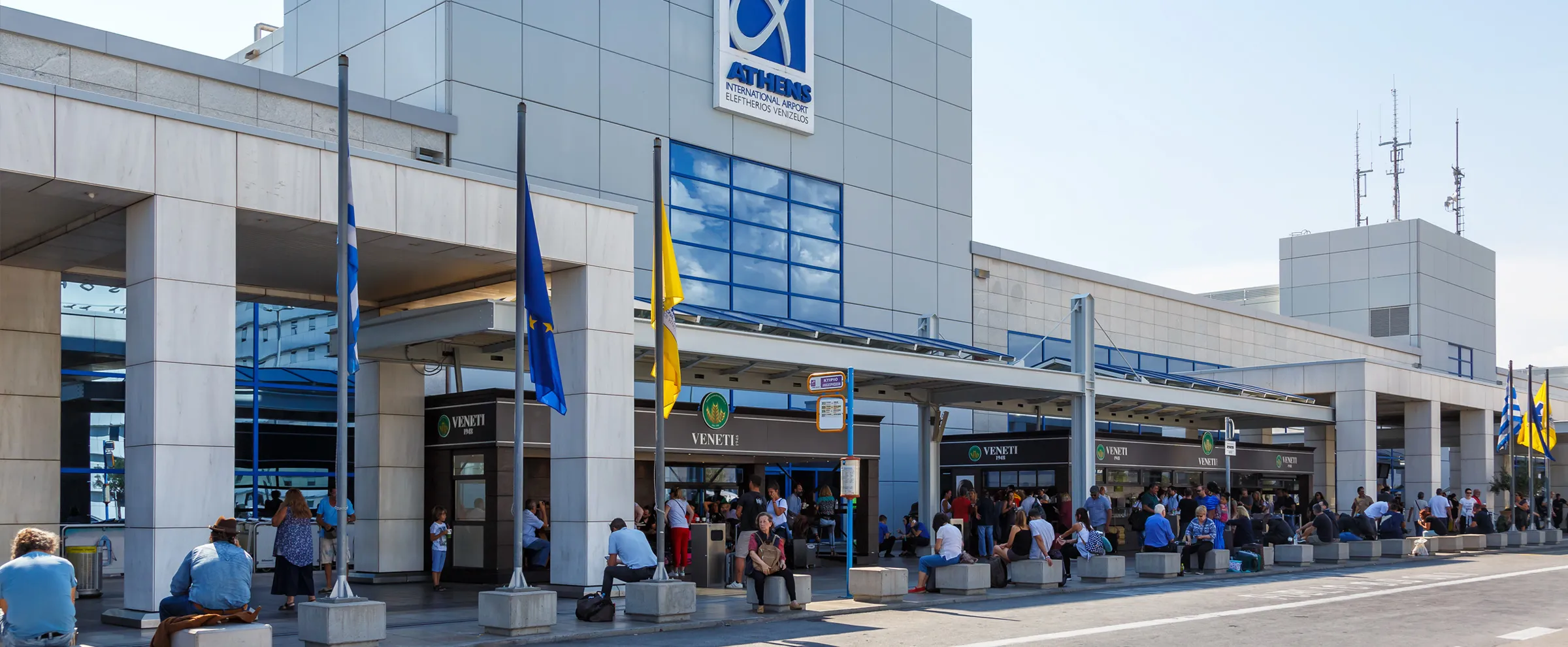 Athens International Airport terminal exterior with people waiting outside and national flags waving.