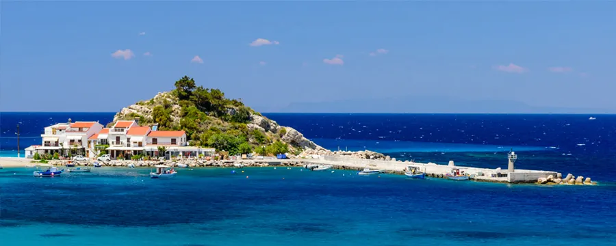 Λιμάνι του νησιού Σάμου με μικρό μόλο, σπίτια και καθαρό γαλάζιο νερό κάτω από φωτεινό ουρανό.