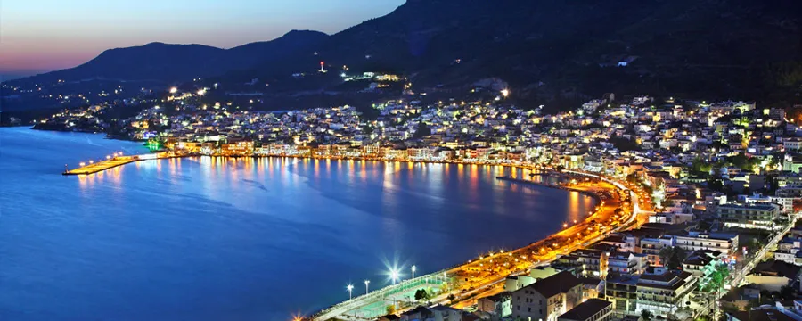 Φωταγωγημένη παραθαλάσσια πόλη της Σάμου τη σούρουπο με ήρεμη θάλασσα και γύρω λόφους στην Ελλάδα.