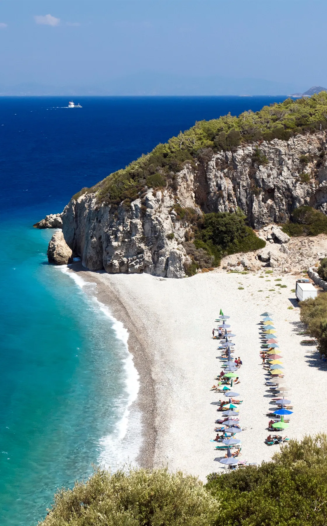 Παραλία στη Σάμο με τιρκουάζ νερό, βοτσαλωτή ακτή, γκρεμό και σειρές ομπρελών και ξαπλώστρων.