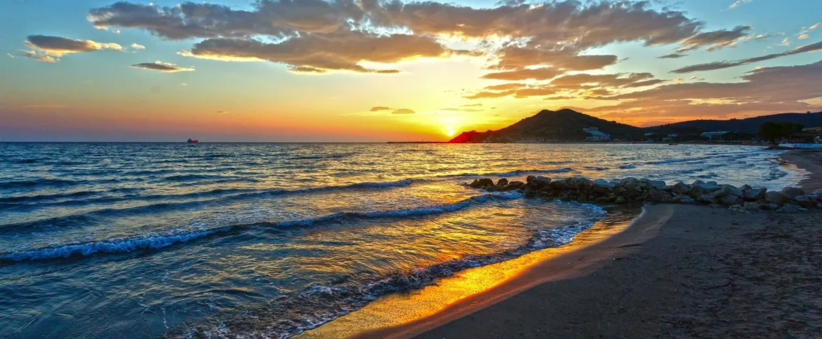 Sunset over Alykes beach in Zakynthos, Greece, with golden waves and a mountainous silhouette.