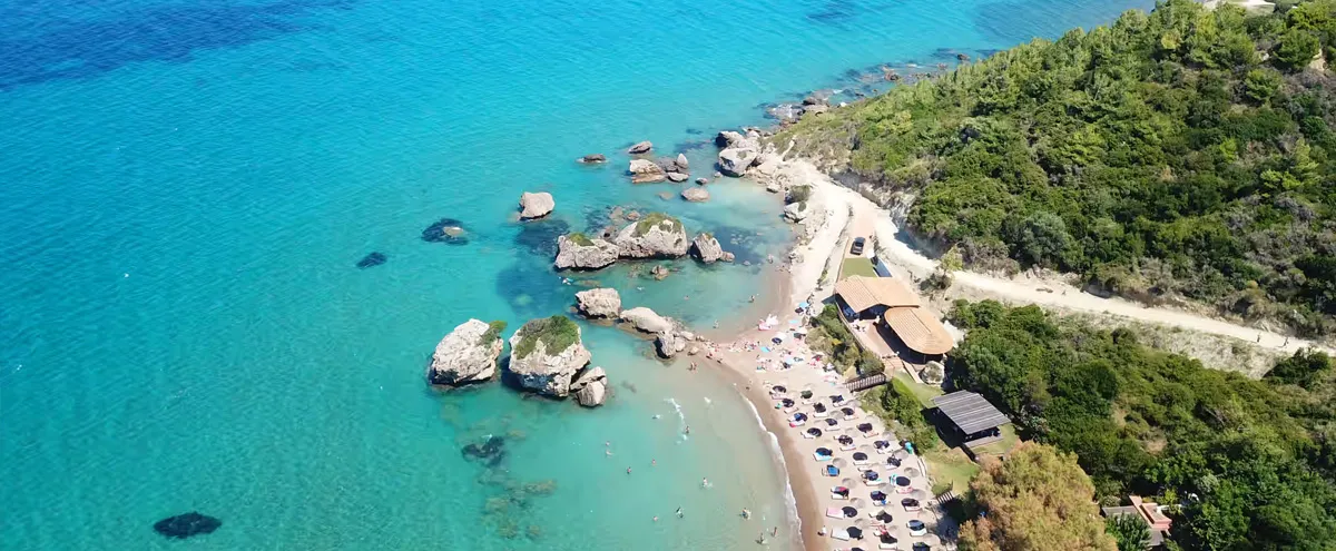 Turquoise waters and rocky coastline surround a sandy beach with sun umbrellas in Vassilikos, Zakynthos, Greece.