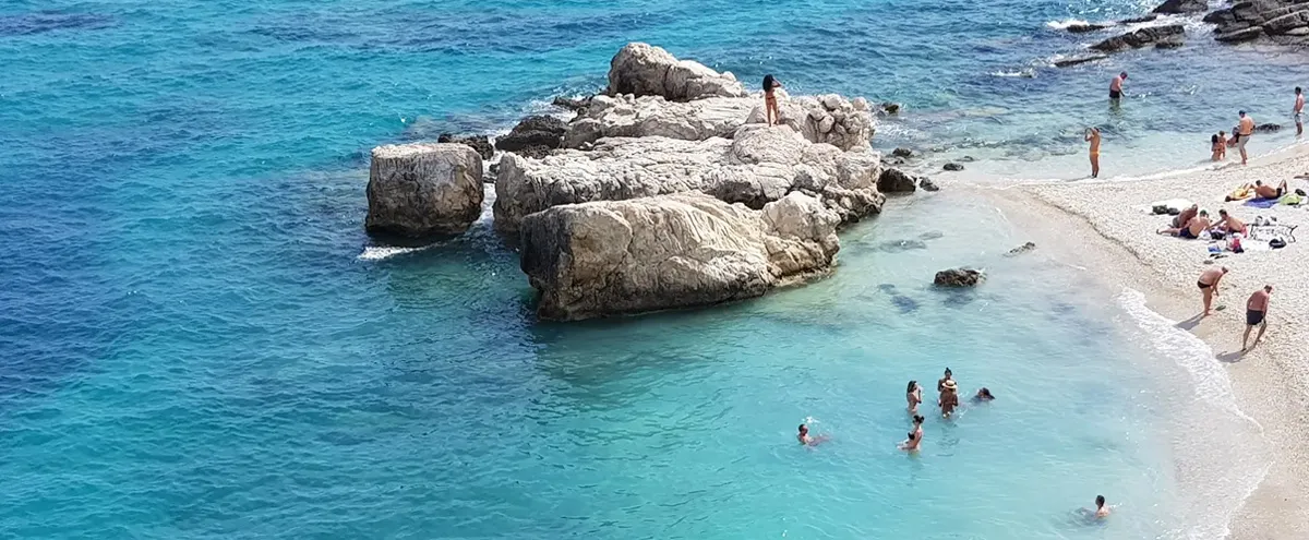 Turquoise waters and large rocks surround swimmers and sunbathers on a sunny Alykanas beach in Zakynthos, Greece.