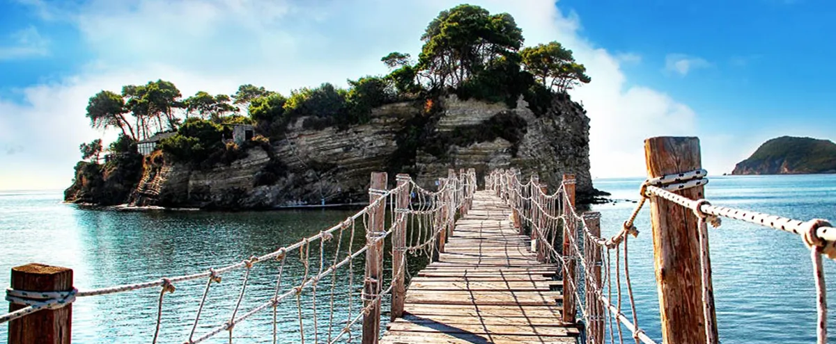 Wooden bridge with rope railings leads to a green, rocky islet surrounded by calm turquoise waters in Zakynthos.
