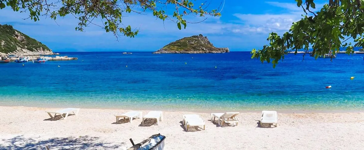 Bright blue sea, sandy beach with sun loungers, green island in the distance at Agios Nikolaos Port, Zakynthos.
