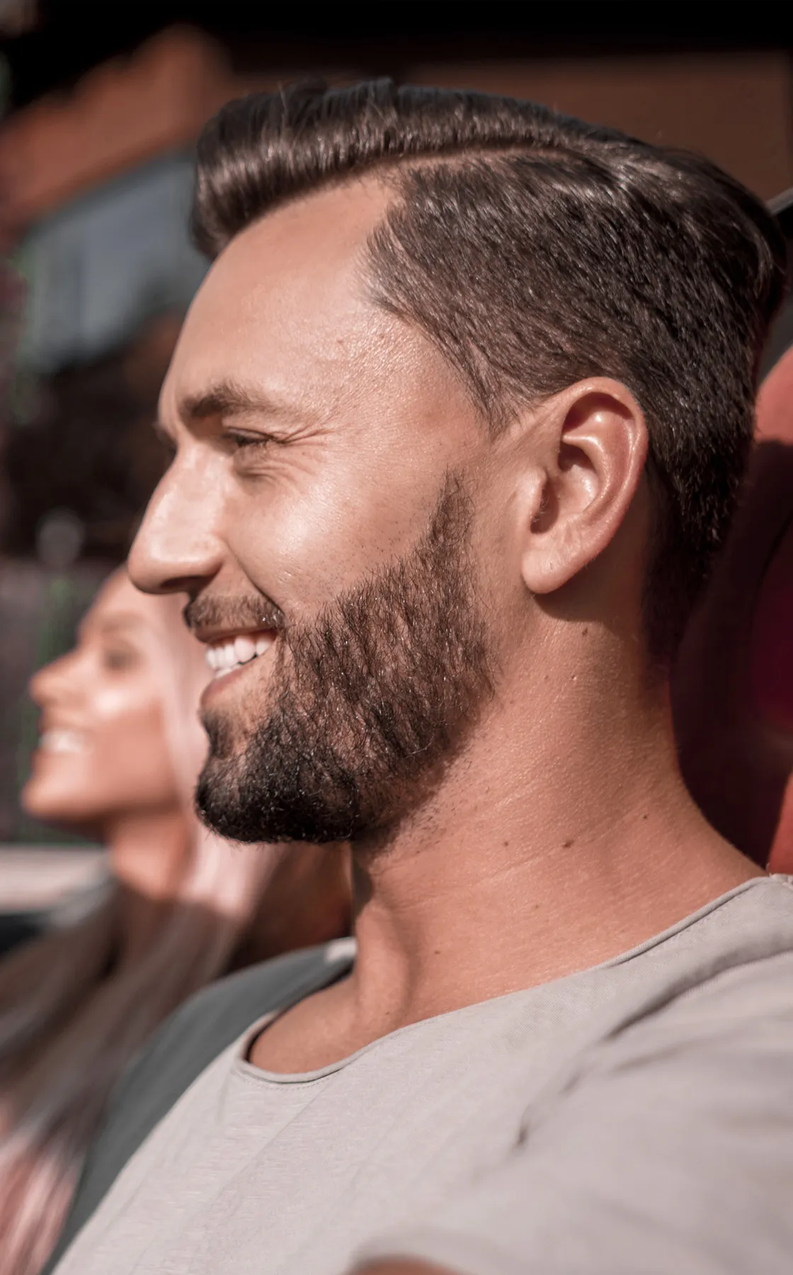 Man met baard en kort haar genietend van zonlicht, vrouw op de achtergrond ontspannen deels zichtbaar.