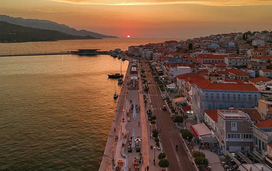 Tramonto dorato sul porto di Samos, con passeggiata affollata e case con tetti rossi lungo la costa.