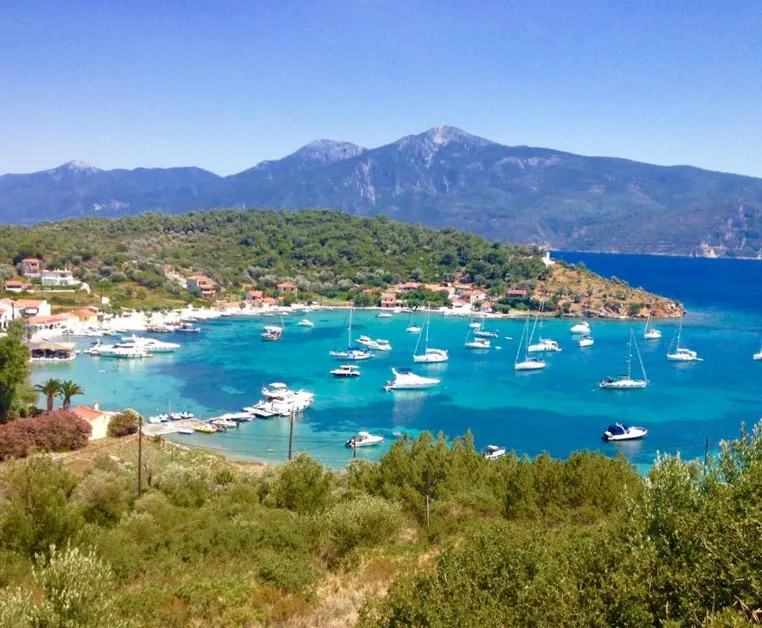 Baia cristallina a Posidonio, Samos, con numerose barche a vela e montagne sullo sfondo soleggiato.
