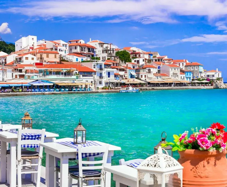 Terrasse en bord de mer avec tables blanches, vue sur les maisons traditionnelles et l’eau turquoise à Kokkari, Samos.