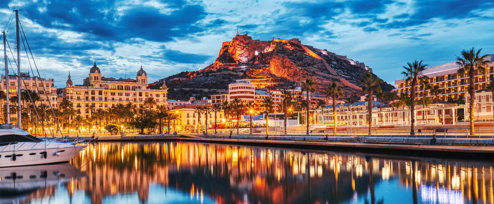 Portul Alicante și castelul Santa Barbara iluminate la amurg cu reflecții în apă.
