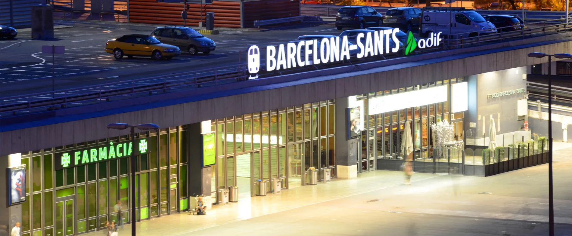 Exterior al stației de tren Barcelona-Sants la amurg, cu farmacie și intrare pentru închiriere auto.