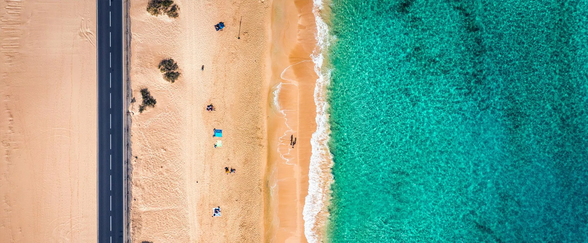 Aerial view of a coastal road next to a sandy beach and turquoise ocean water on a clear day.