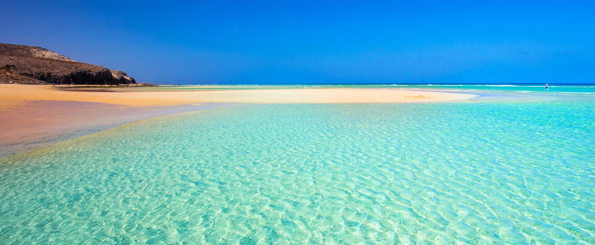 Crystal clear shallow water and sandy beach with rocky hills under a bright blue sky.