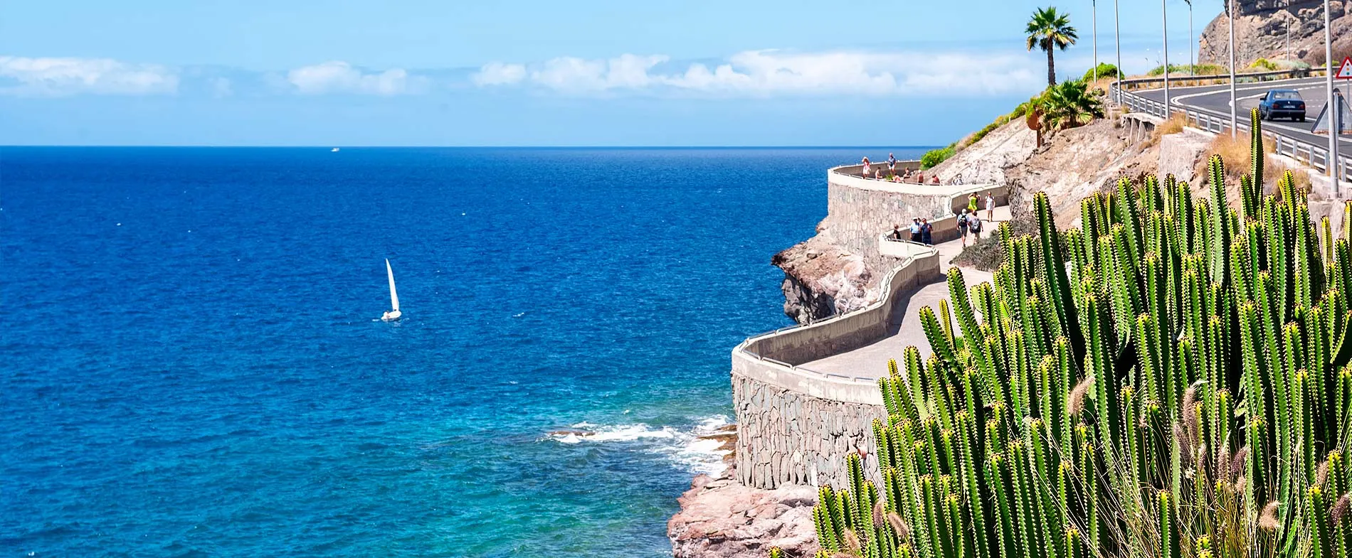 Strada costiera e percorso pedonale con persone che ammirano l'oceano blu e una piccola barca a vela.
