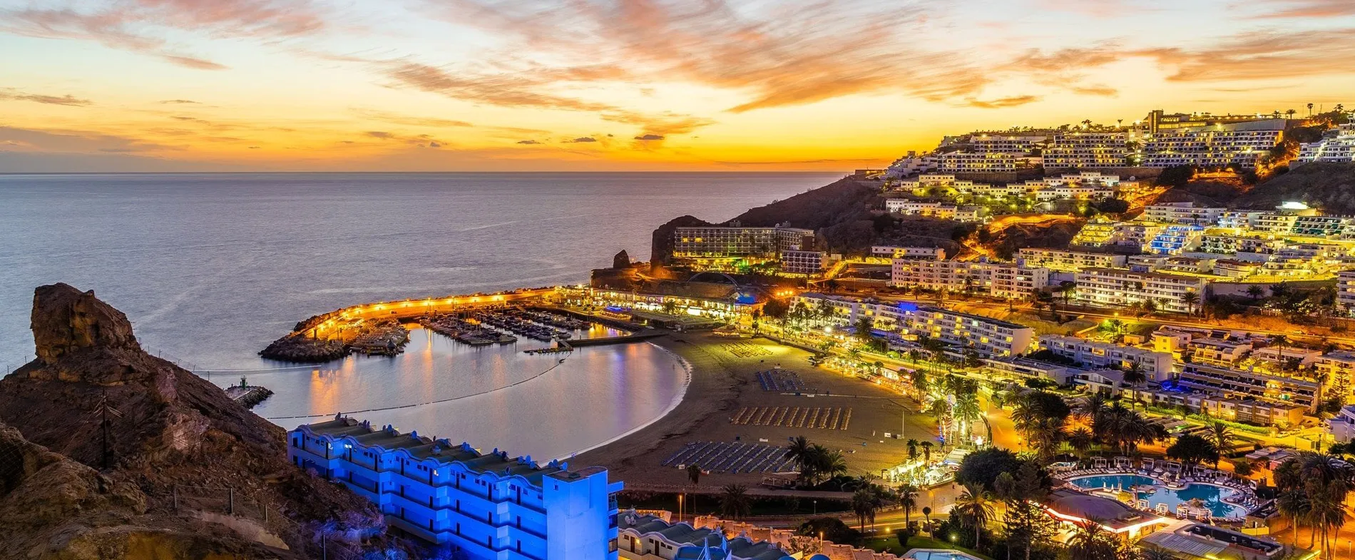 Sunset view over a coastal resort town with a marina, beach, and hillside buildings lit up at dusk.
