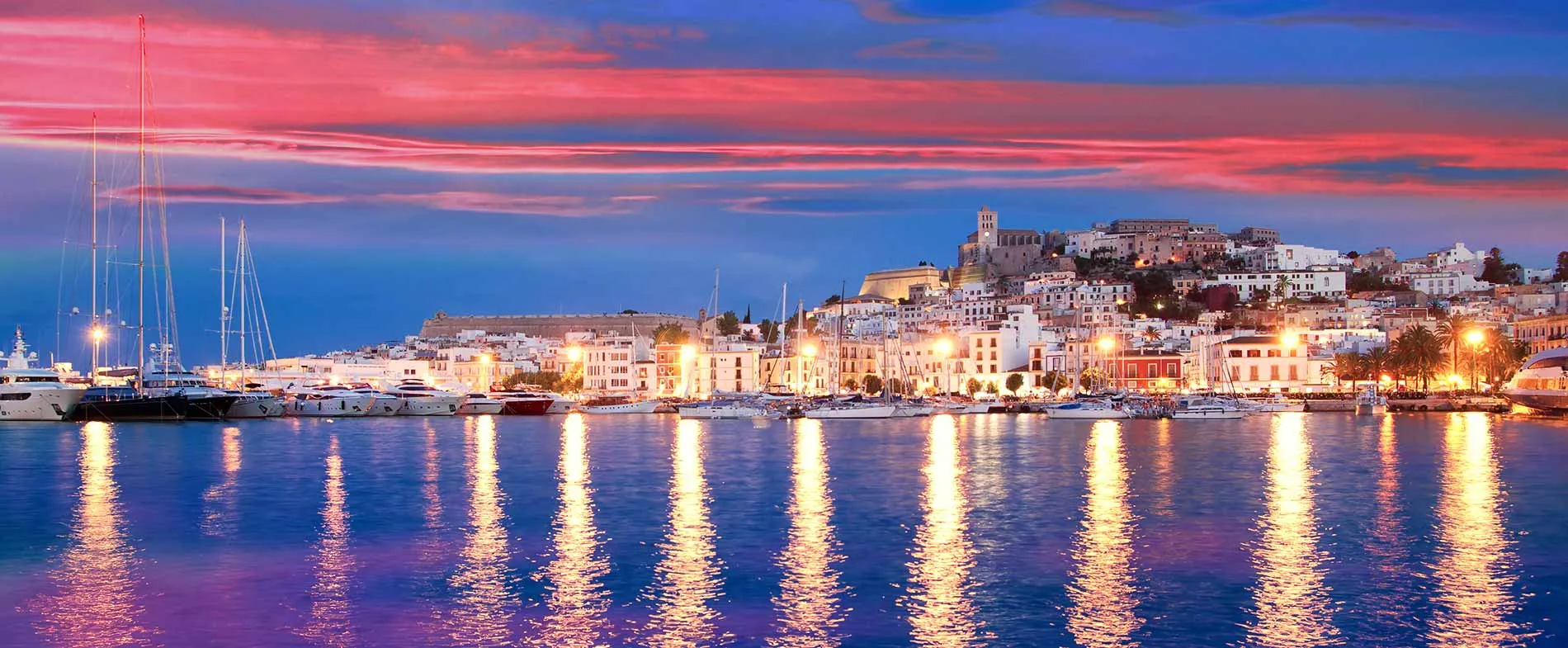 Ibiza Hafen bei Sonnenuntergang mit Yachten und Stadtlichtern im Wasser.