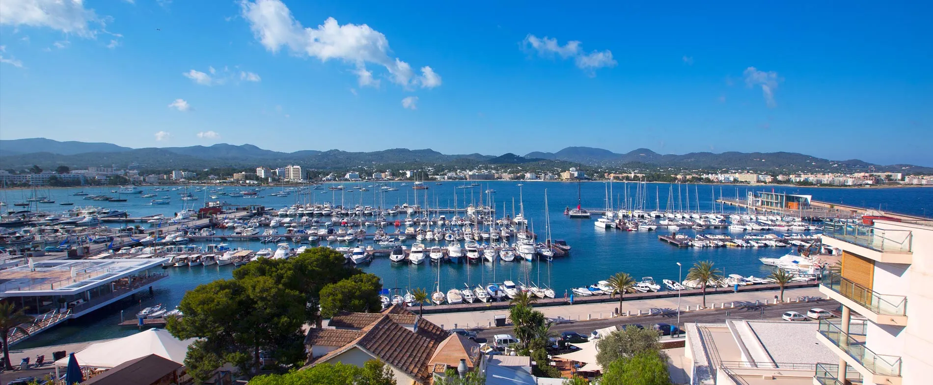 Ibiza marina with boats docked and cityscape nestled by hills in the background on a clear day.