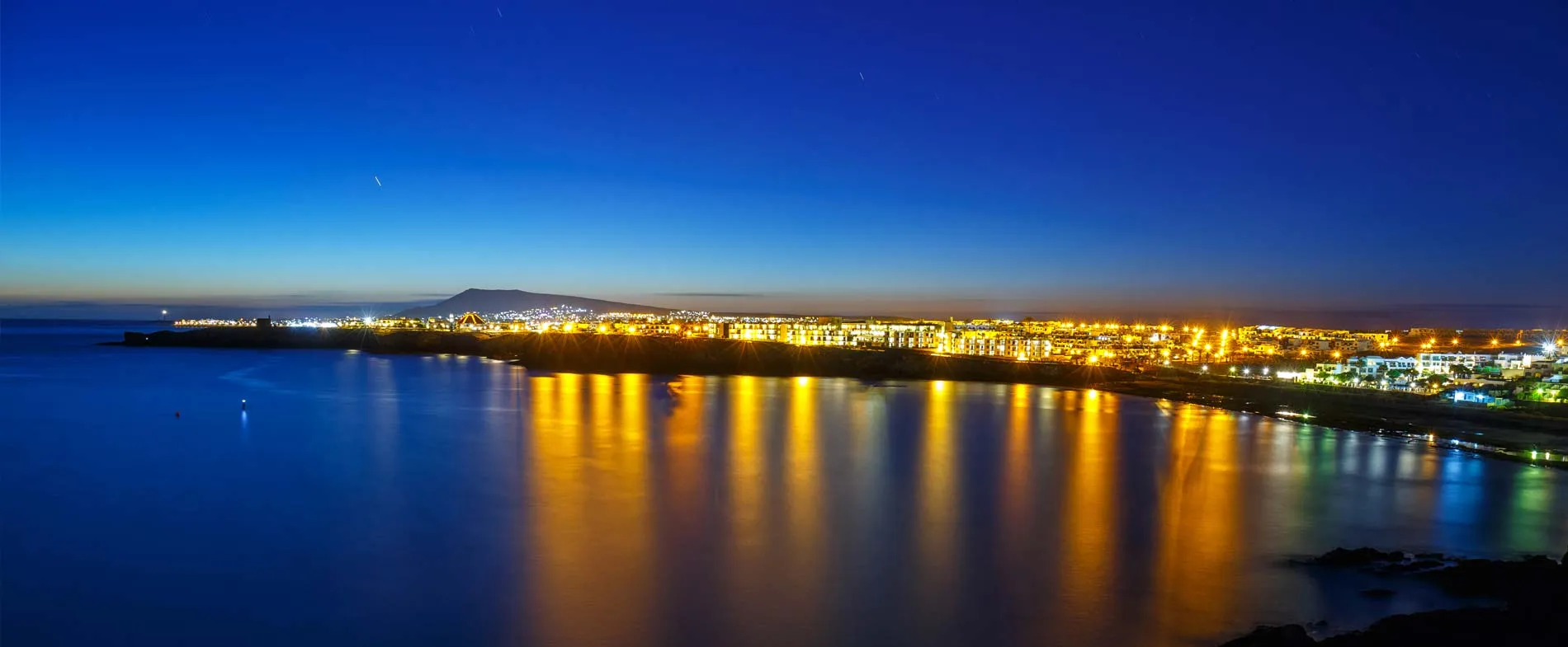 Peisaj urban de coastă la amurg, cu apă calmă reflectând luminile puternice ale orașului.