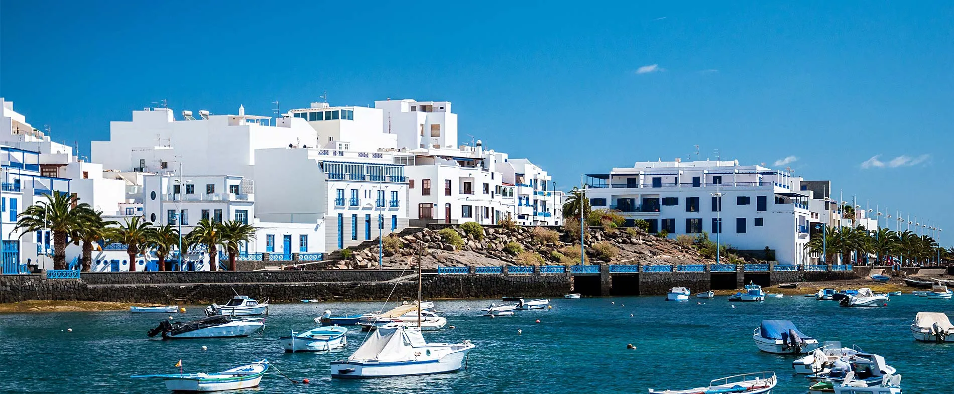 Coastal town with white buildings and small boats moored along the calm waterfront under clear blue skies.