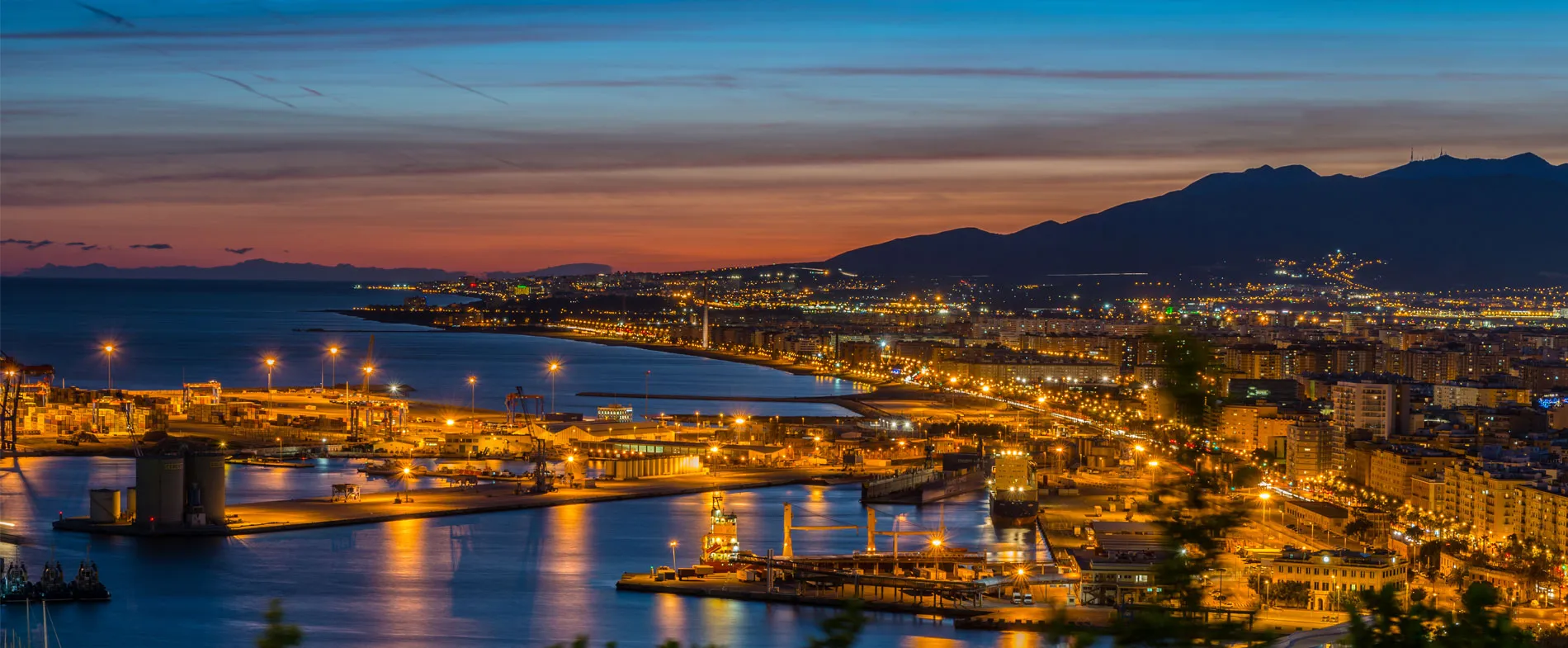 Coasta Malagă la amurg cu luminile orașului și port, ideal pentru închiriere de mașini.