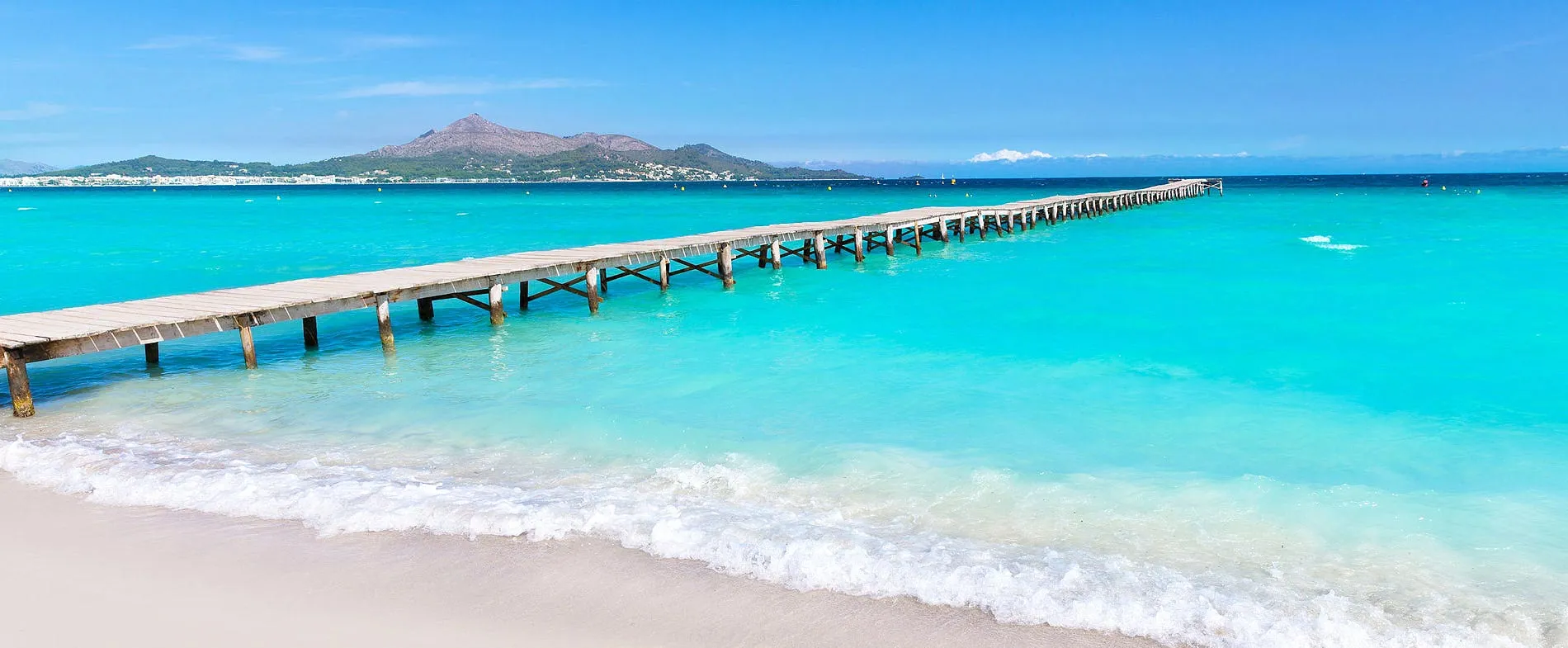 Molo di legno che si estende sulle acque turchesi della spiaggia di Alcudia con montagne sullo sfondo.