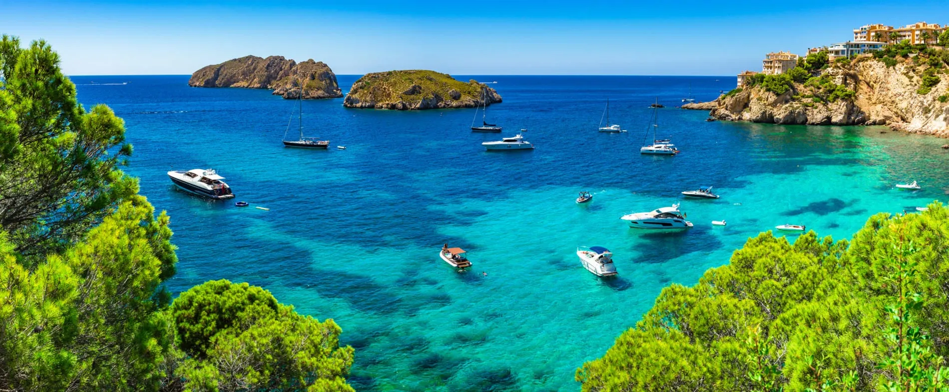 Clear blue sea with sailboats and yachts near rocky islands and pine-covered shores in a sunny Mediterranean bay.