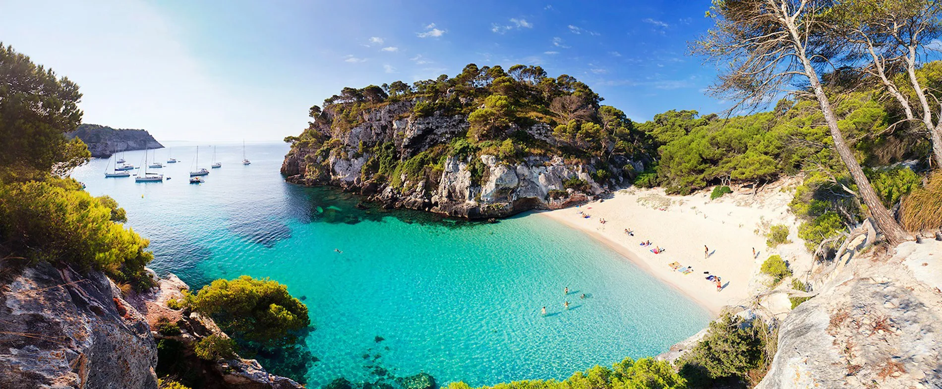 Cala Macarelleta beach in Menorca with turquoise water, sandy shore, rocky cliffs, and boats anchored nearby.