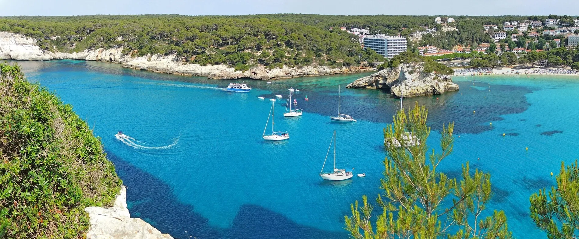 Menorca coastline with boats and a sandy beach surrounded by pine trees and rocky cliffs.