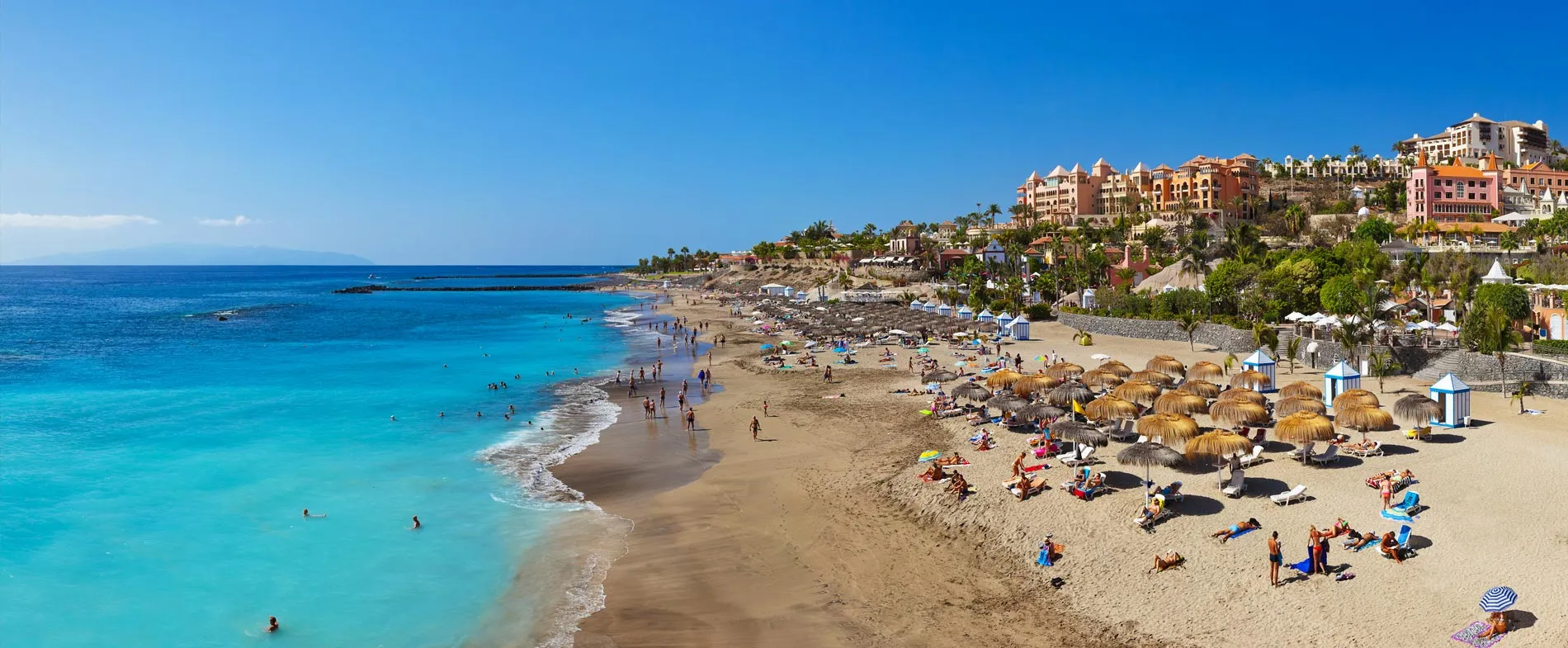 Tenerife beach with clear blue water and sunbathers enjoying a sunny day near resort buildings.