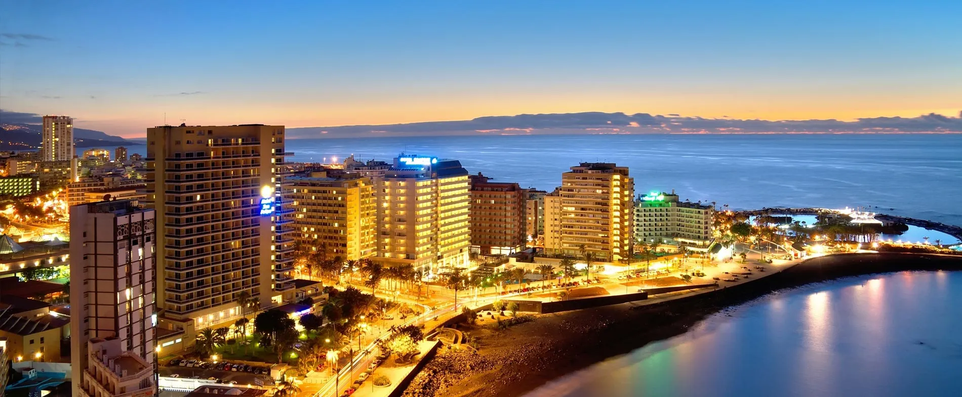Vedere costieră a orașului Tenerife la amurg cu clădiri luminate și apă liniștită pentru închiriere auto.