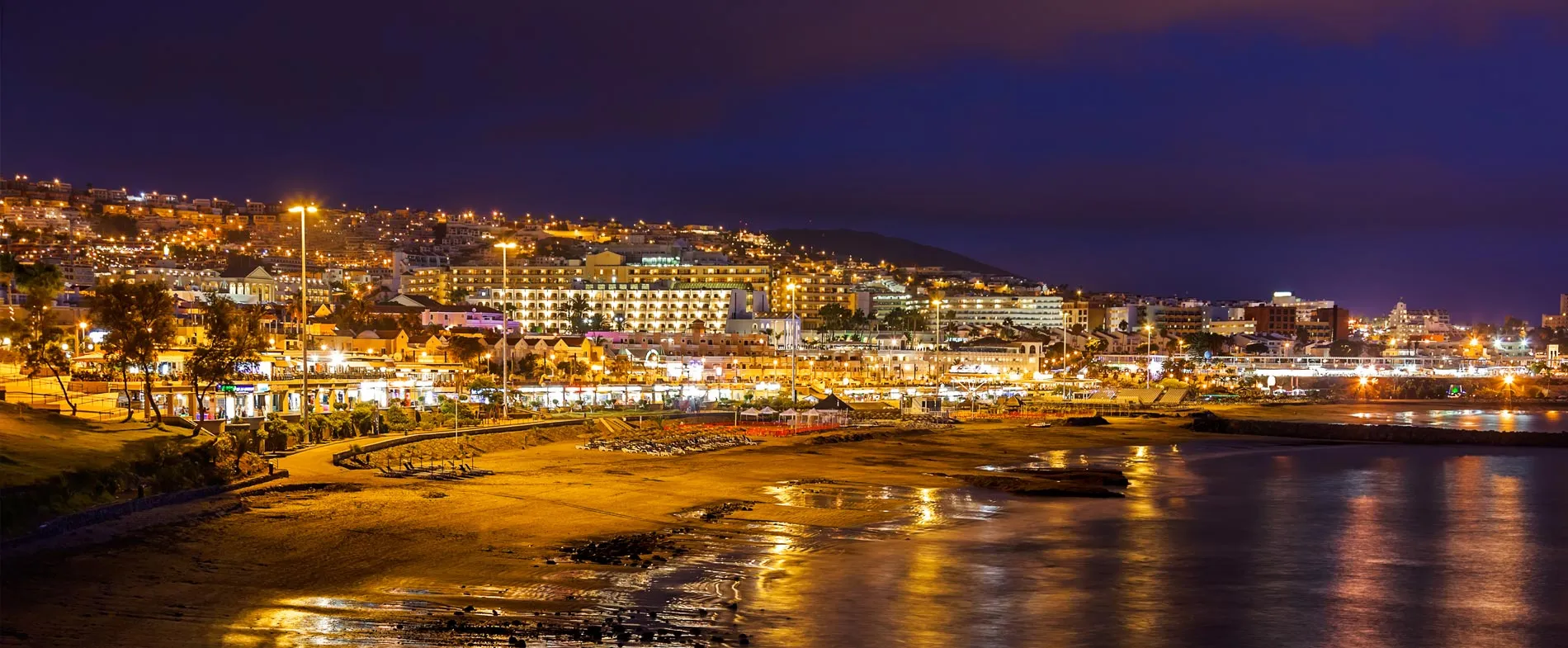 Vedere de noapte a unui oraș coastal iluminat cu clădiri și plajă care reflectă luminile pe apă.