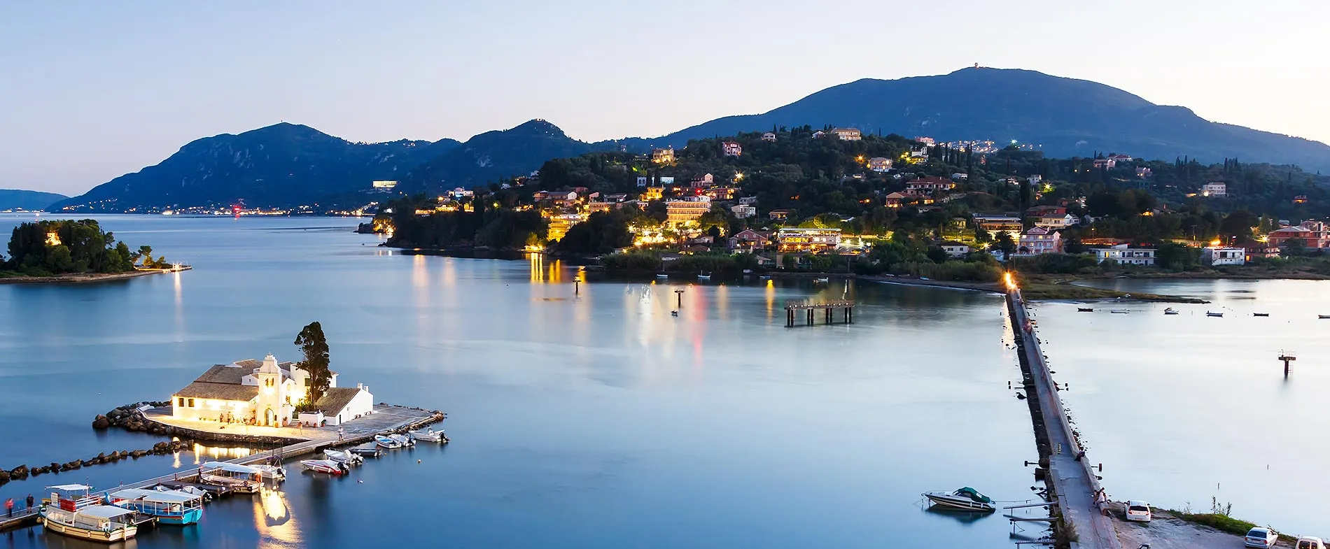 Costa dell'isola di Corfù con edifici illuminati e una piccola chiesa vicino al mare calmo al tramonto.