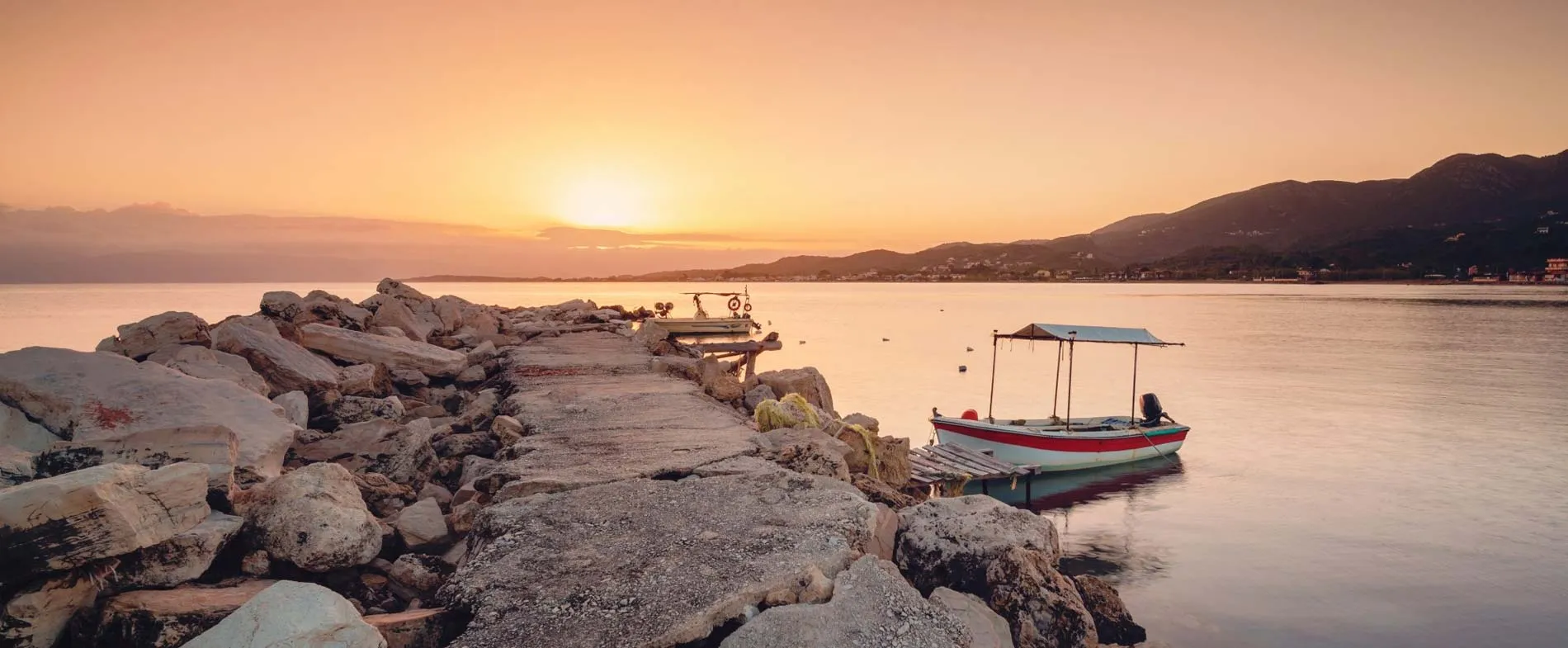 Tramonto su un molo roccioso con una piccola barca da pesca attraccata su acque calme vicino alle colline della costa.