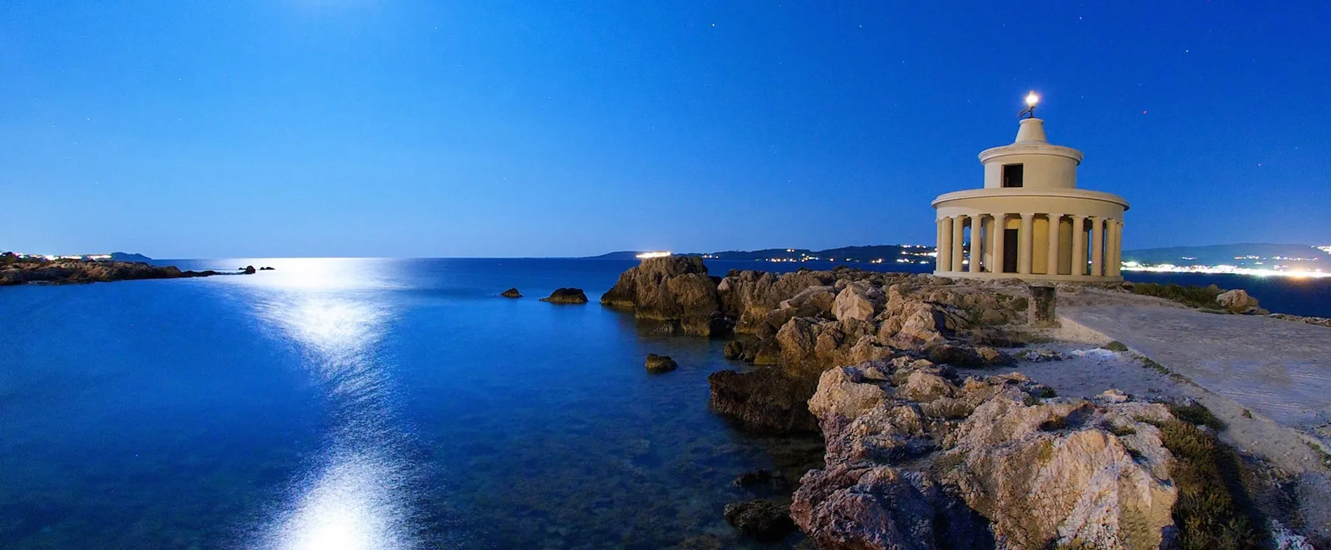 Lighthouse on rocky coast of Kefalos, Kos, lit under a clear night sky by the calm sea.