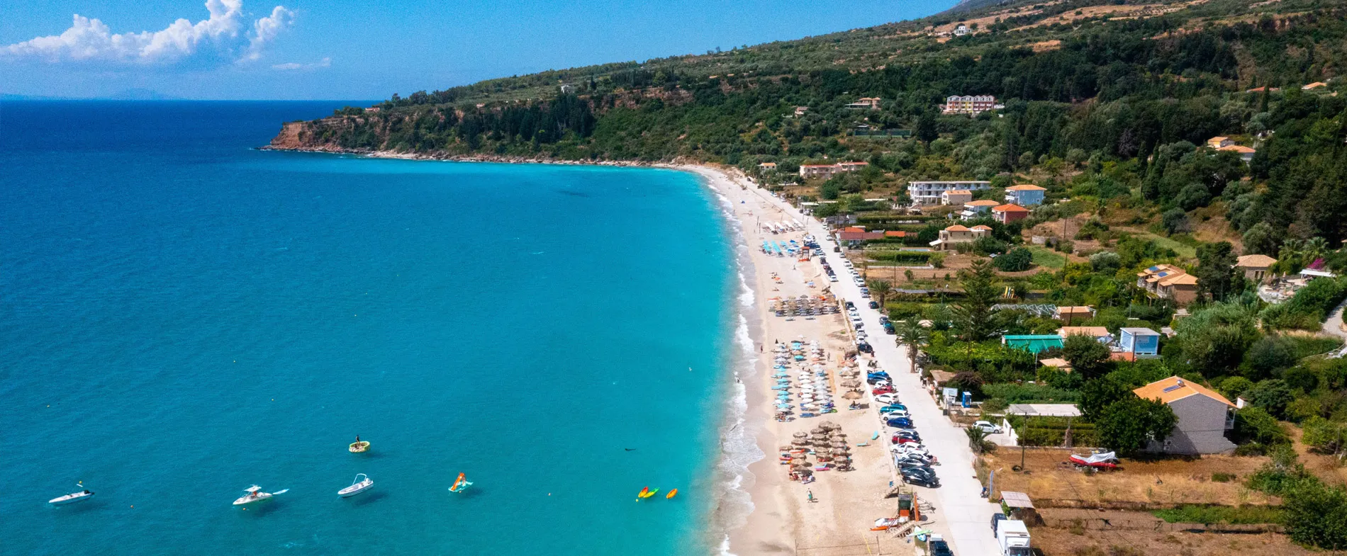 Aerial view of a sandy beach with umbrellas, boats on clear blue water, and green hillside with houses nearby.