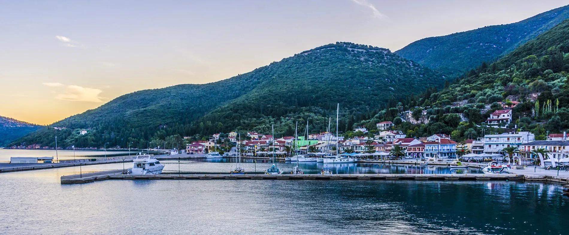 Coastal town port with boats docked and green hills in the background at sunset.