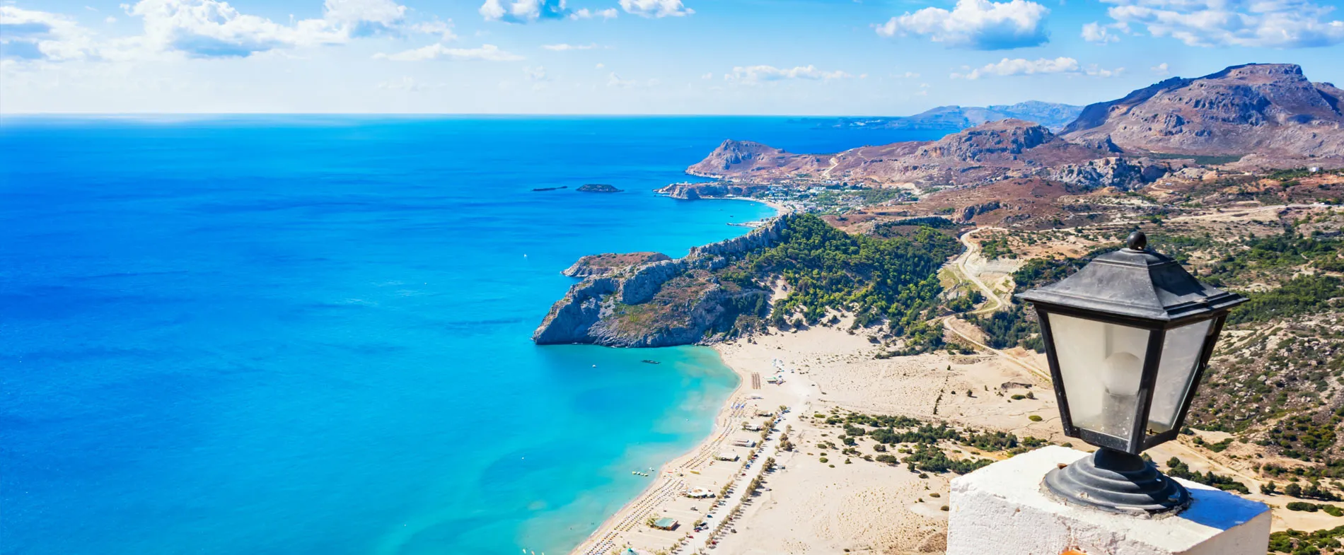 Rhodes coastline with clear blue sea, sandy beach, and rocky hills under a partly cloudy sky.