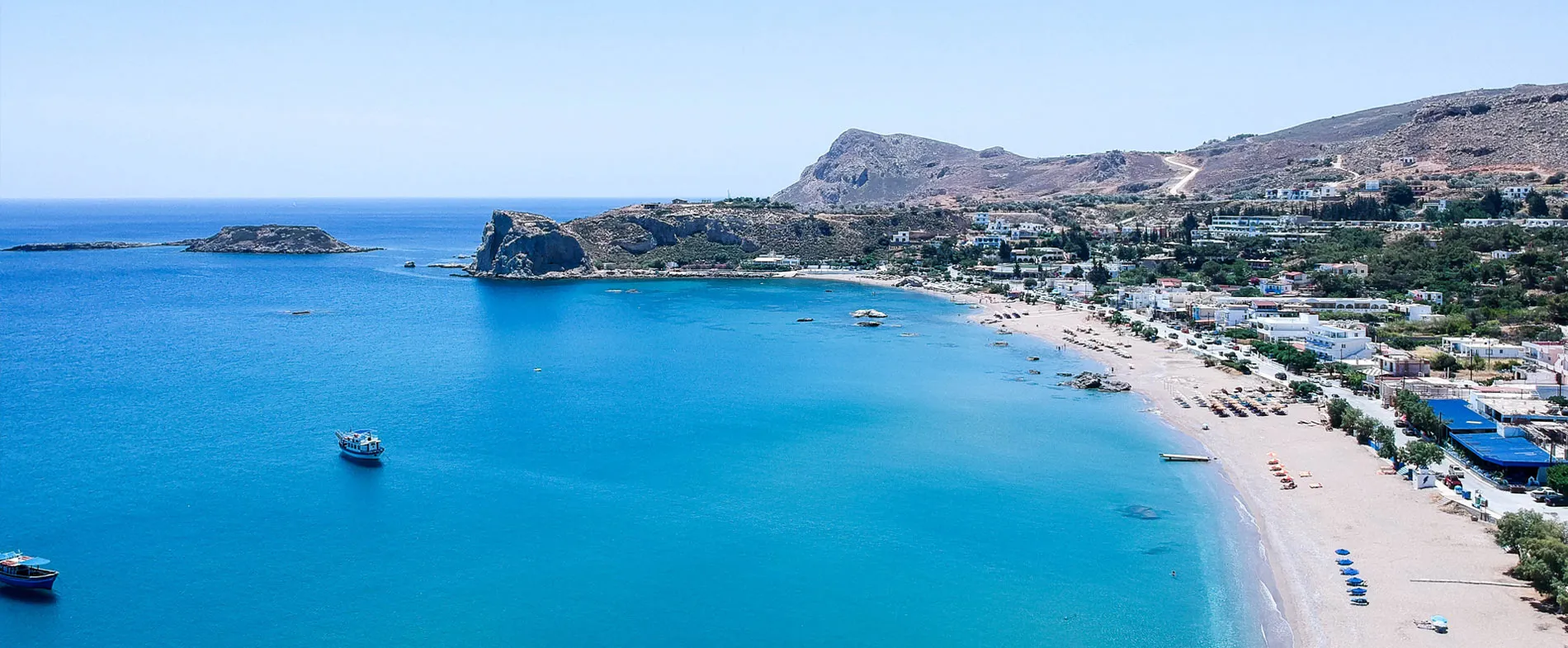 Rhodes island coastline with clear blue sea, boats, sandy beach, and hillside village buildings.