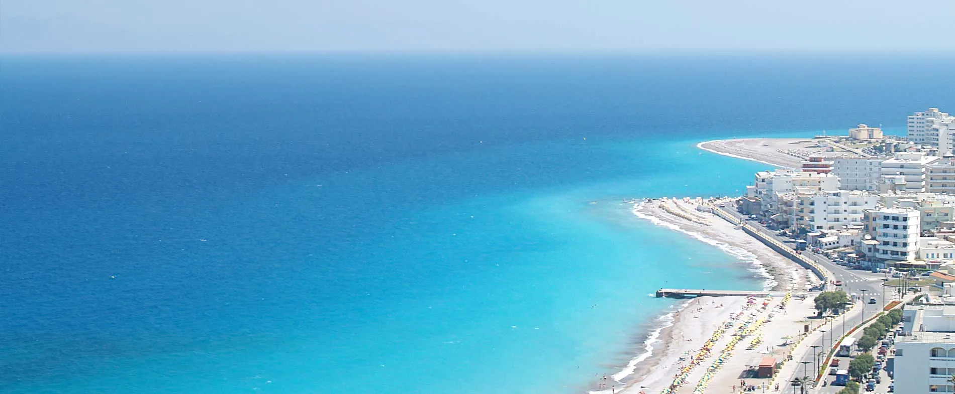 Coastal view of Rhodes showing clear blue sea and beaches lined with sun umbrellas near buildings.