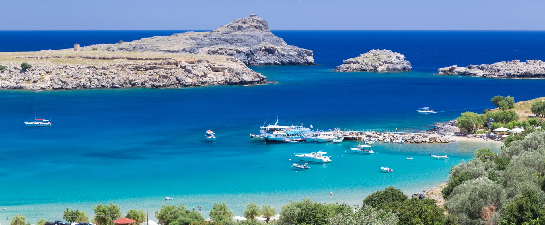 Rhodes bay with turquoise waters, boats anchored, rocky islands and a small beach with sun umbrellas.