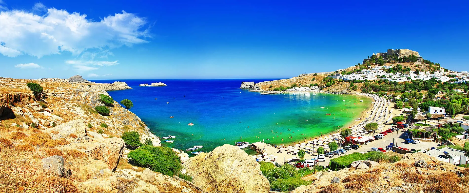 Lindos Bay in Rhodes with turquoise sea, sandy beach, sun umbrellas, and hillside village with fortress.