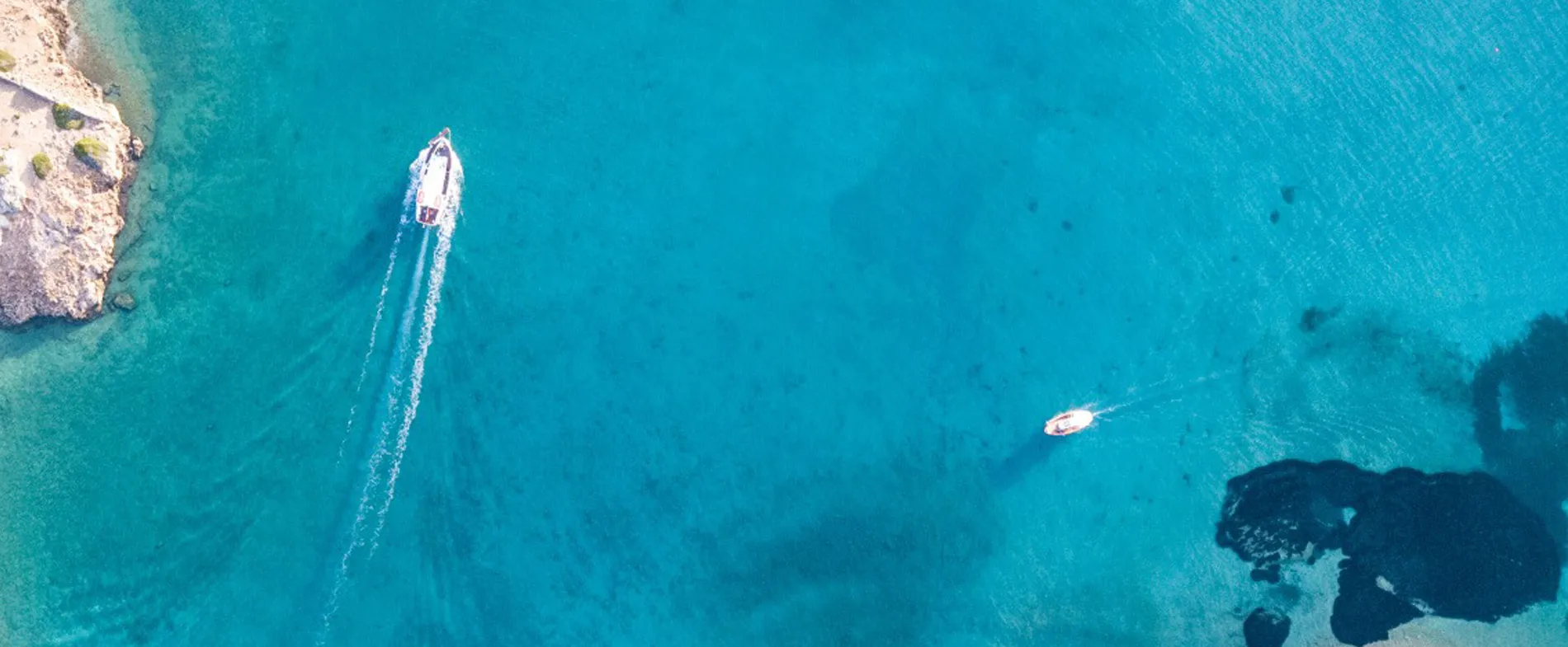 Due barche navigano su acqua azzurra limpida vicino a una costa rocciosa sotto un sole luminoso.