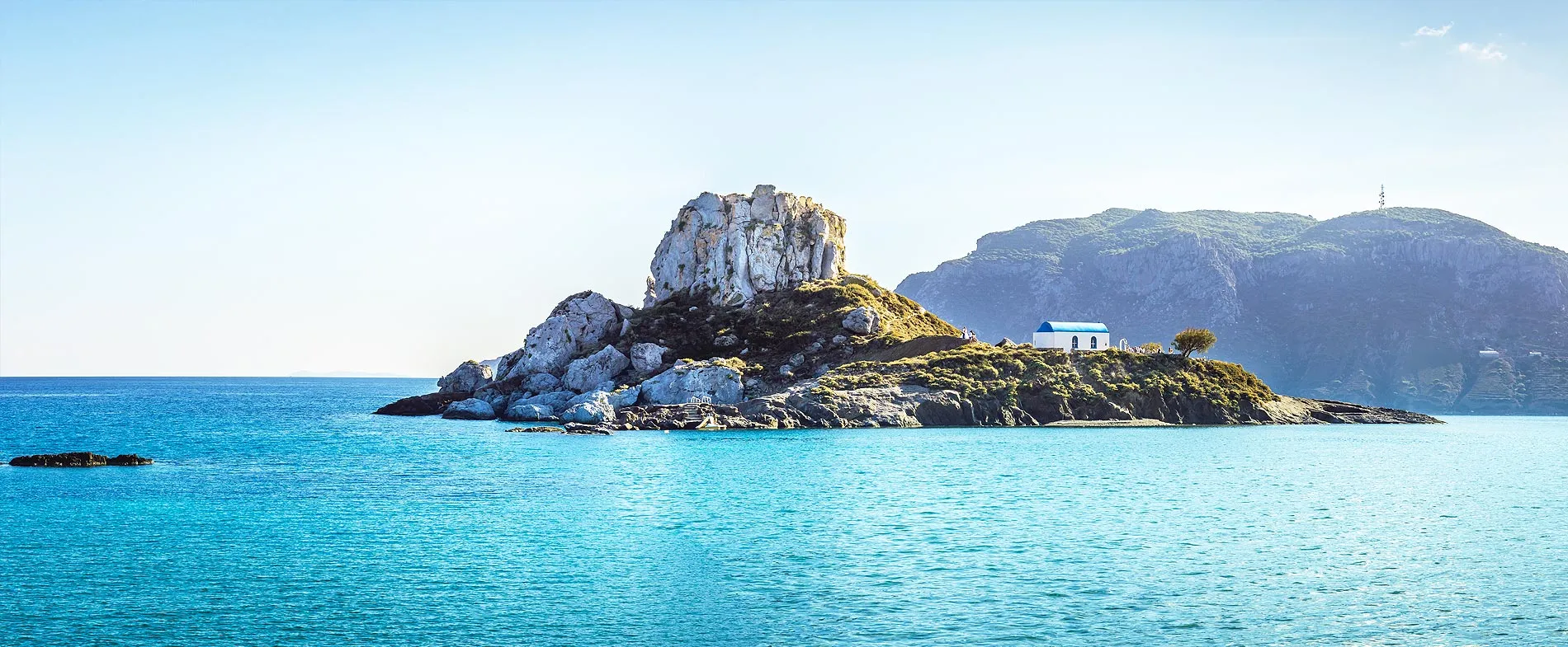 Piccola isola rocciosa con cappella bianca dal tetto blu, circondata da mare azzurro e montagne lontane.