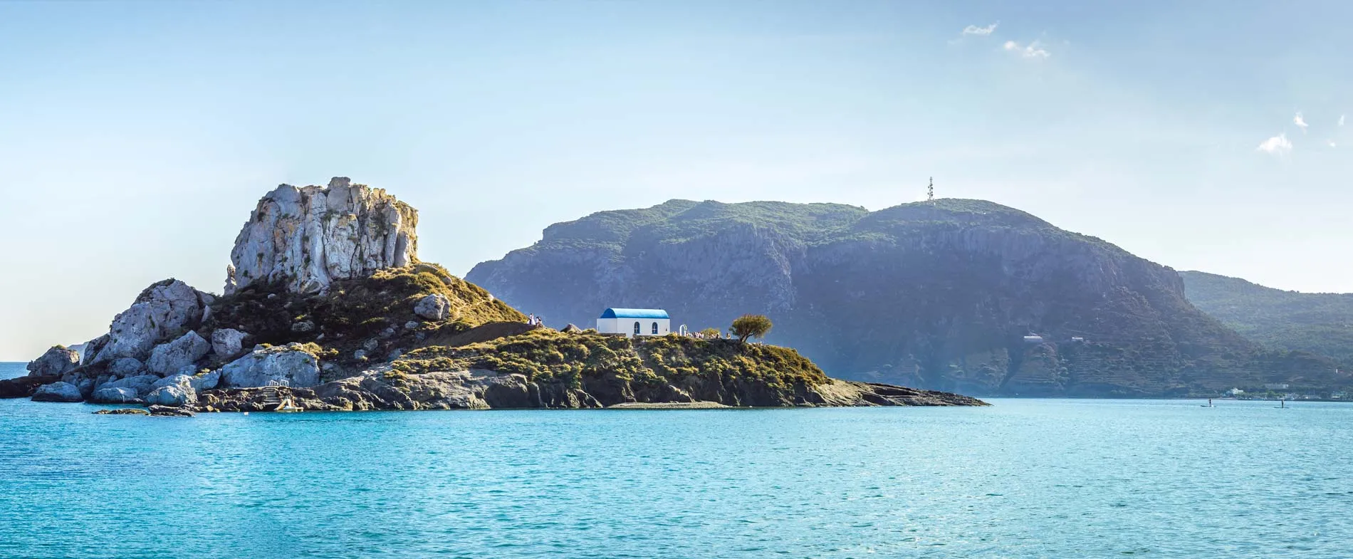 Small rocky island with a white chapel and blue roof near Kos Island, Greece, surrounded by clear turquoise sea.