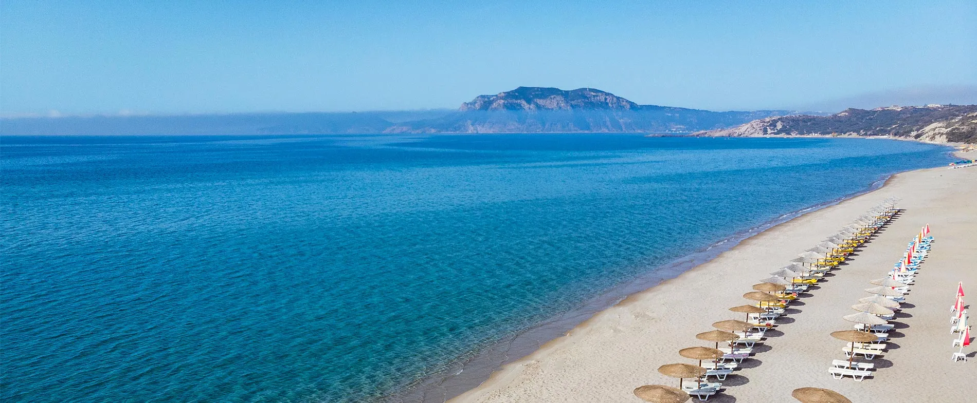 Long sandy beach with rows of sunbeds and umbrellas by clear blue sea and distant mountains.