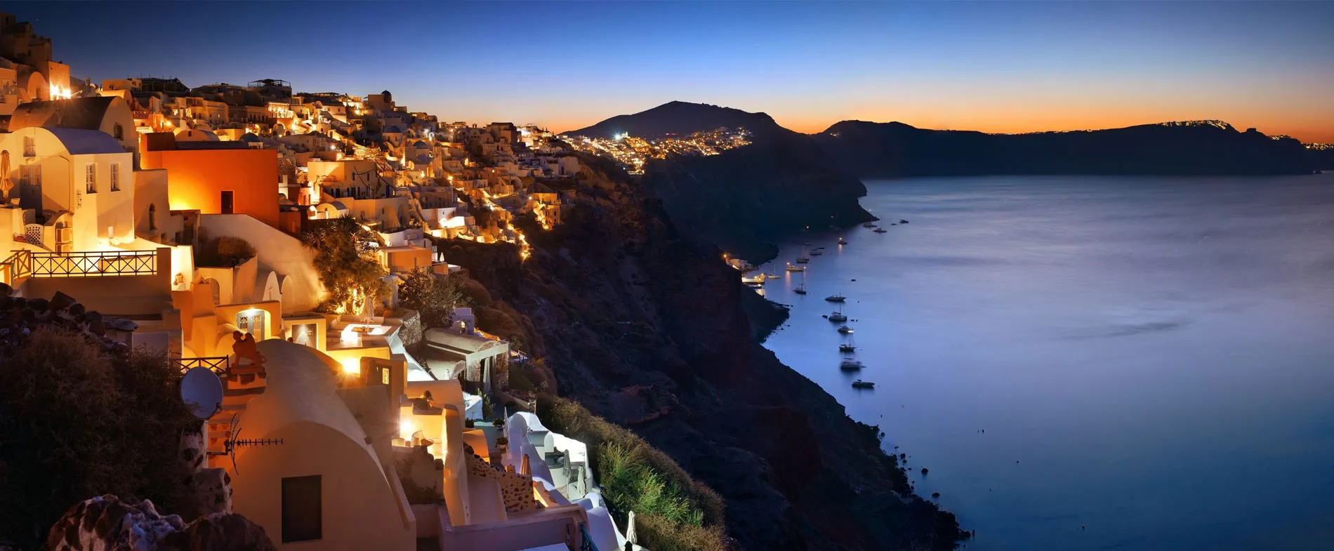 Santorini coastline with illuminated white buildings by the sea at dusk for car rental vacations.