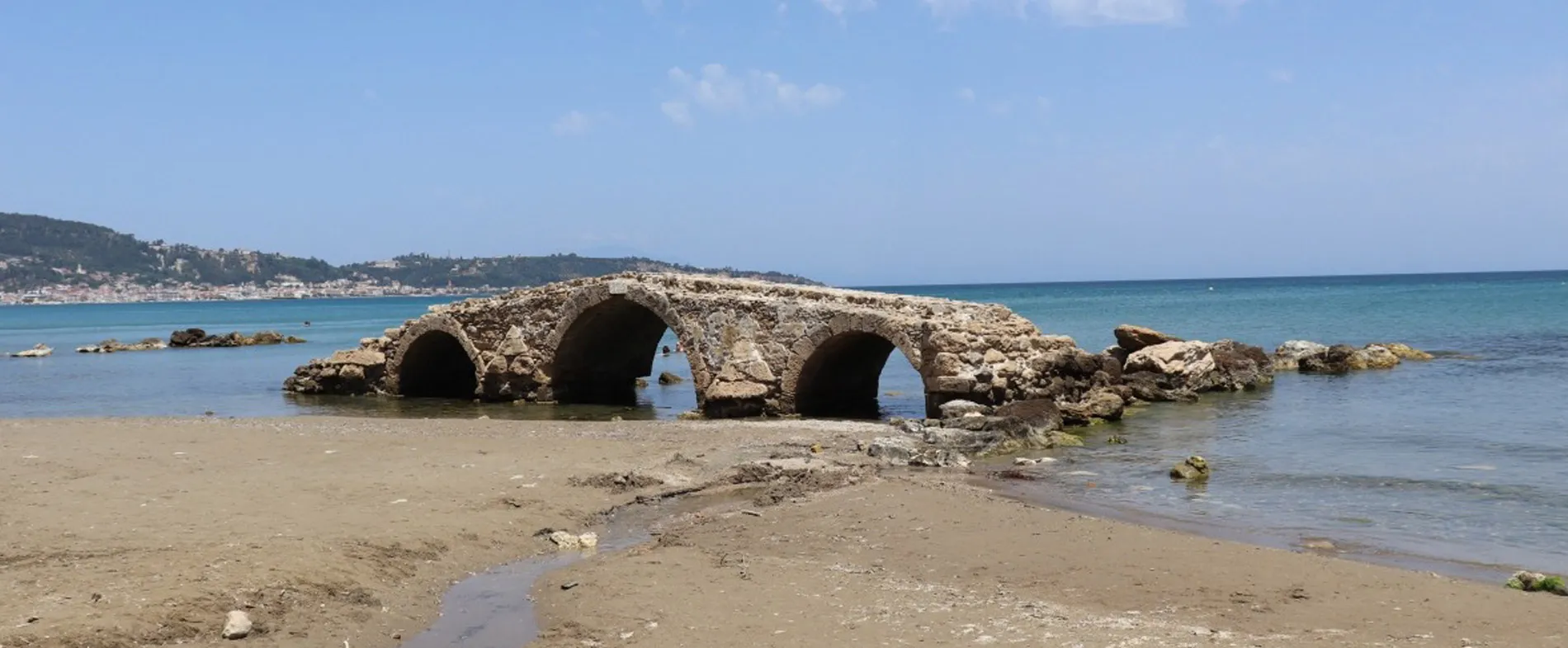 Ancient stone bridge over shallow water on a sandy beach with blue sea and clear sky.